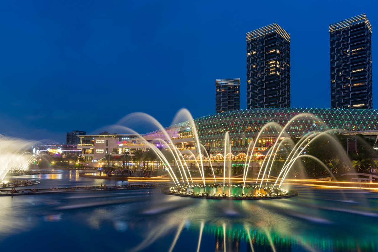 A large fountain with three tiers is illuminated by lights, creating a mesmerizing display of water sprays. The fountain is surrounded by a circular pool, and the background features tall buildings with reflective surfaces. The scene is set against a dark blue sky, emphasizing the fountain's vibrant colors.