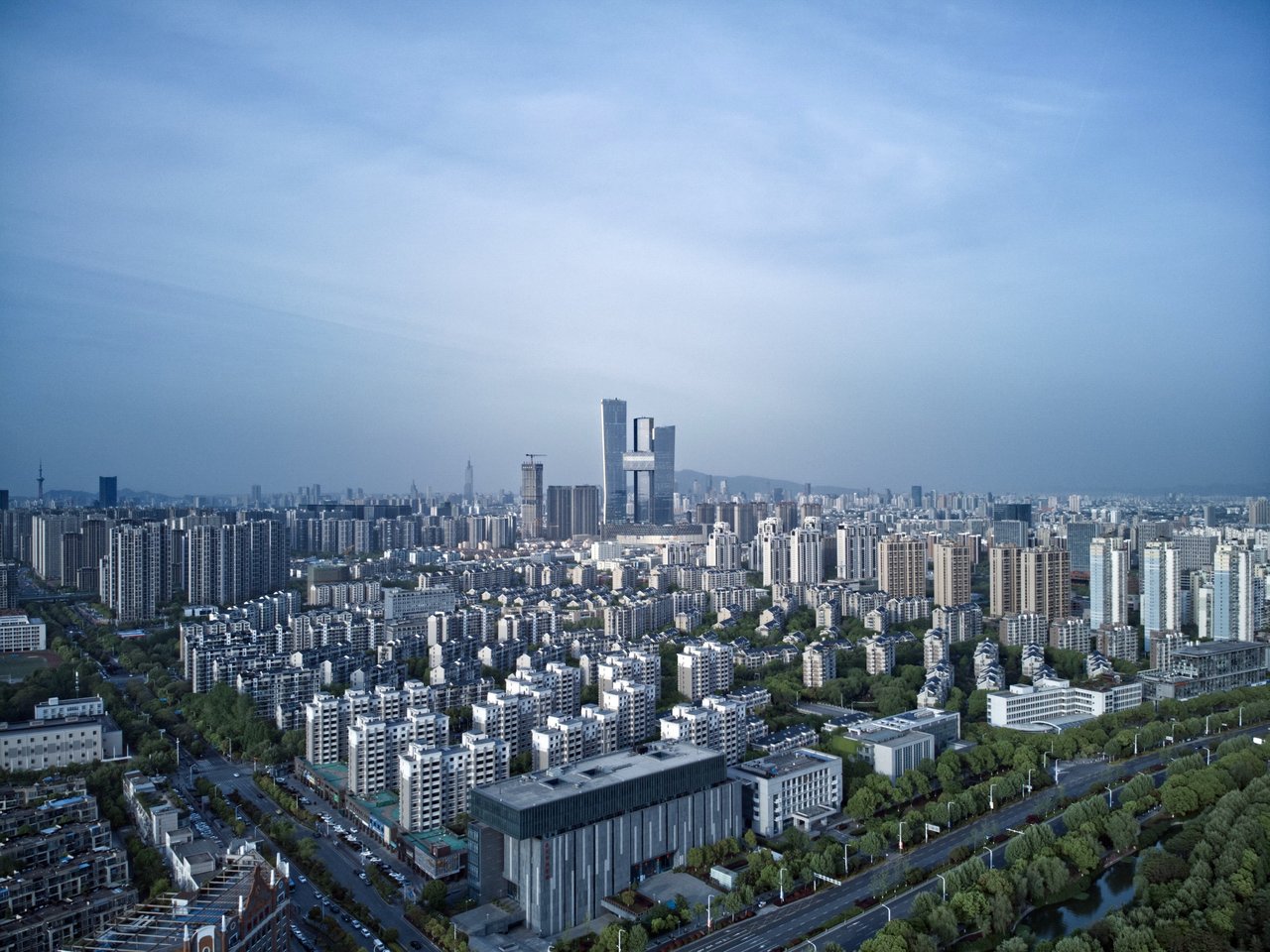 A bustling cityscape with numerous tall buildings, including skyscrapers and a tower, is captured from a high vantage point, showcasing the urban landscape's density and architectural diversity.