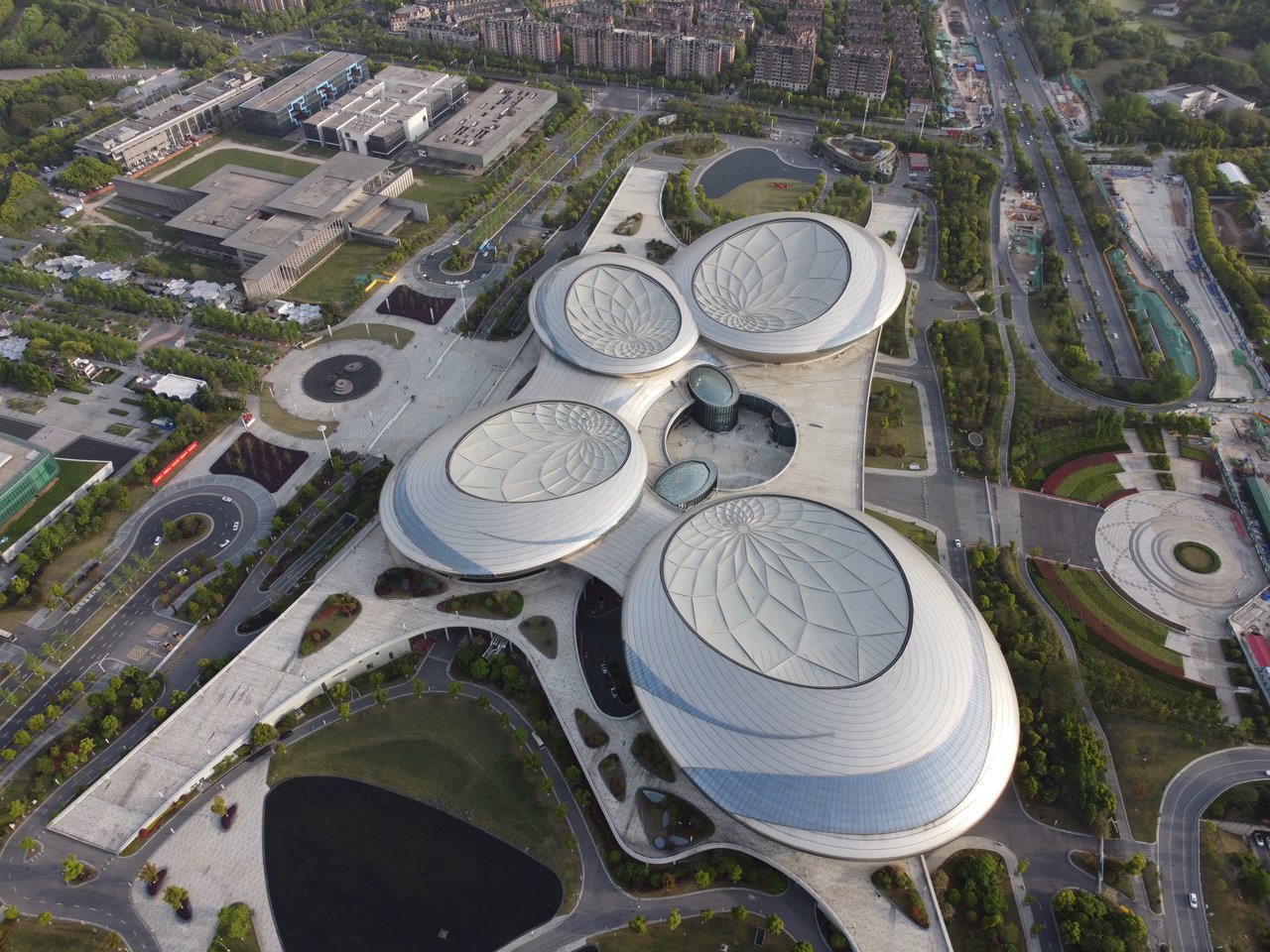 An aerial view of a modern, large, circular building surrounded by a park and a road, with a large circular pool in the center.