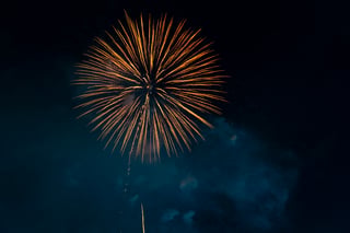 A large, bright orange and yellow firework explosion fills the night sky, with a streak of light extending from the top left corner. The explosion is centrally located, surrounded by dark clouds.