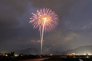 A fireworks display illuminates the night sky, with a large explosion in the center and smaller ones scattered around. The cityscape below is illuminated by lights, and mountains are visible in the distance.