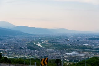 A city with a winding river, mountains, and a mountain range in the background, with a road and a yellow and black traffic sign in the foreground.