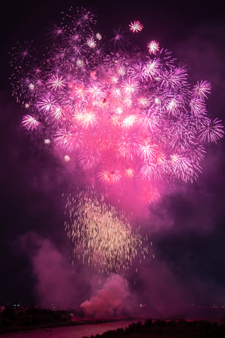 A vibrant fireworks display illuminates the night sky, with pink and purple hues and a mix of white and yellow streaks. The fireworks are scattered across the sky, with some closer to the viewer and others further away. The water below is calm, and a few boats are visible in the distance.
