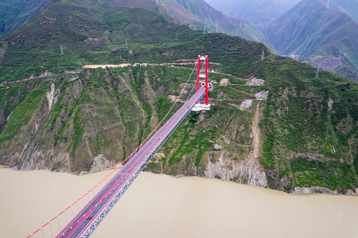 A red and white suspension bridge spans a river, with two red towers on either side. The bridge is surrounded by green mountains and a cloudy sky.