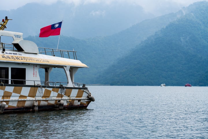 A large white boat with a checkered pattern, a red and yellow flag, and a yellow railing is on calm water, with a smaller boat in the distance. The scene is set against a backdrop of lush green mountains and a cloudy sky.