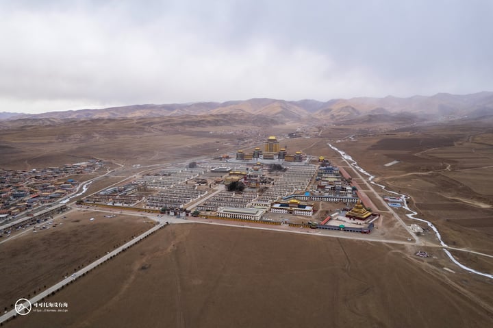 An aerial view of a large, complex construction site surrounded by barren mountains and a cloudy sky. The site features a mix of buildings, roads, and equipment, with a prominent golden dome.