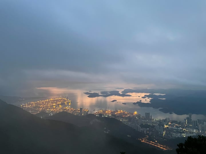 A foggy cityscape is seen from a high vantage point, with a large body of water in the foreground and a mountain range in the background.