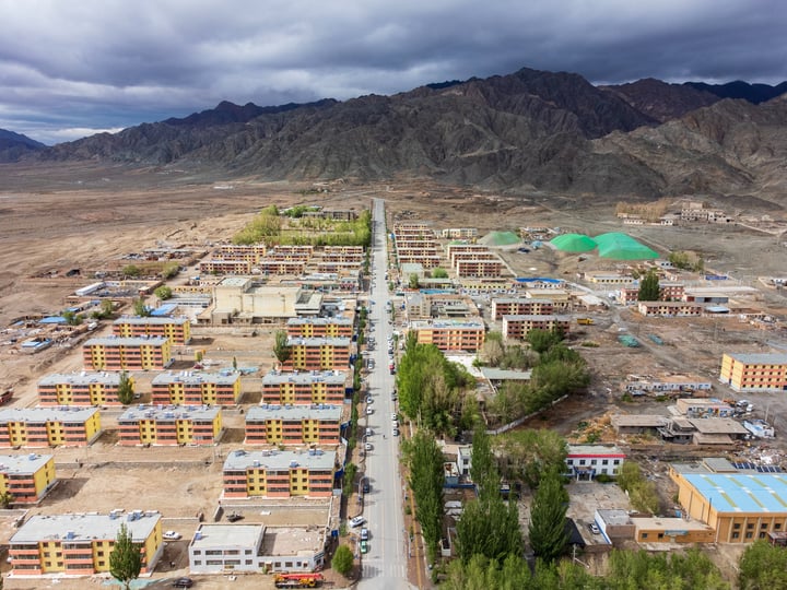 An aerial view of a mountainous area with a road, buildings, and green spaces, with a cloudy sky overhead.