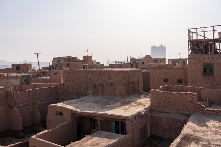 A low-angle view of a desert cityscape with brick buildings, a tall skyscraper in the distance, and a few scattered trees.