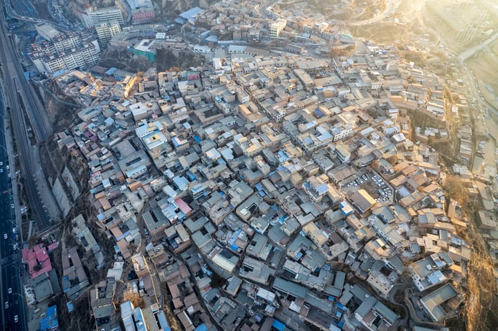 An aerial view of a densely populated city with a mix of white and gray buildings, some with blue roofs, surrounded by roads and highways. The sun casts a warm glow, highlighting the city's architectural details and the surrounding landscape.