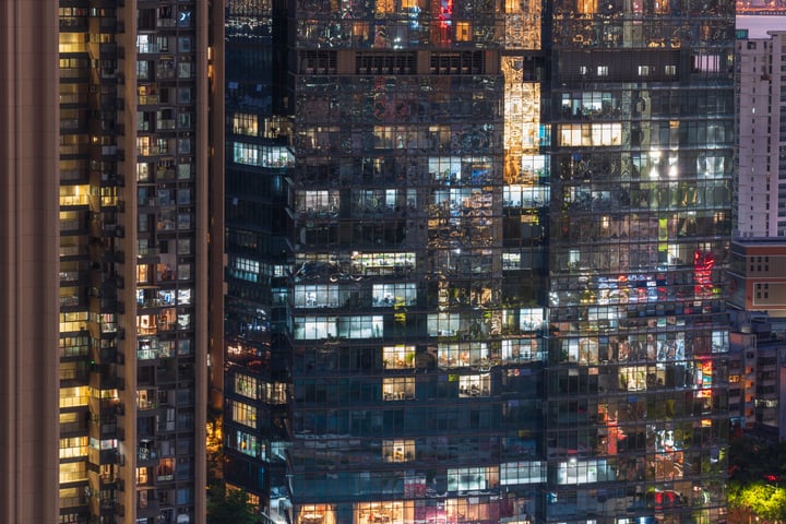 A nighttime cityscape features tall, reflective buildings with lit windows, reflecting the city's lights and creating a mesmerizing effect. The buildings are densely packed, with some having multiple levels, and are illuminated by various lights. The scene is set against a dark blue sky, and the image is taken from a high vantage point, emphasizing the city's grandeur and architectural complexity.