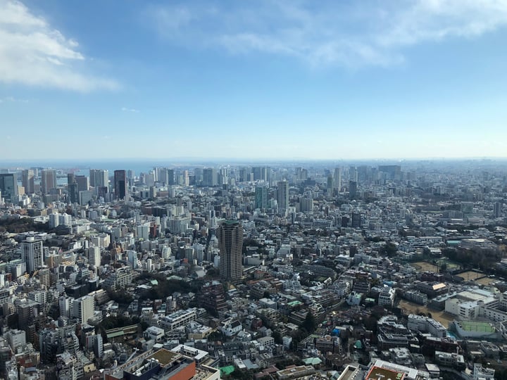 A bustling cityscape with towering skyscrapers and a clear blue sky, with a few scattered trees and a few cars on the streets.