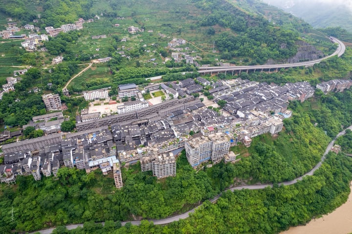 An aerial view of a mountain town with gray and brown buildings, winding roads, and a river, surrounded by lush greenery.