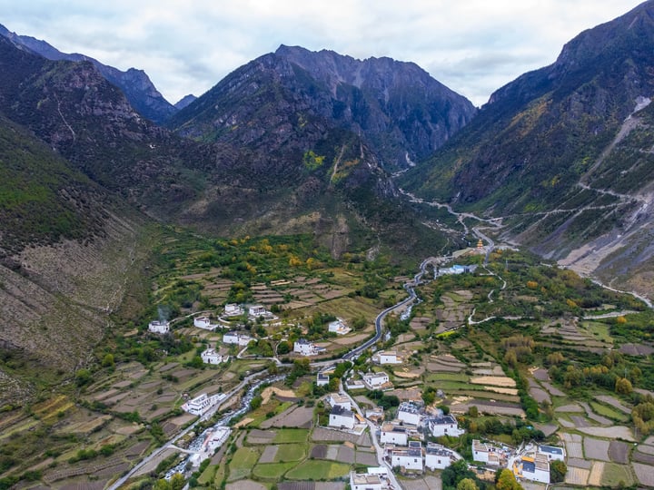 An aerial view of a valley with a winding river, surrounded by mountains, with white houses and green fields, and a cloudy sky above.