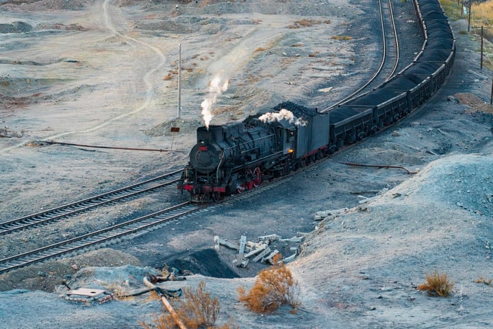 A black steam locomotive with a red front and black cars travels along a curved track in a rugged, rocky landscape. The train is surrounded by rocks and dirt, and the sky is a pale blue.