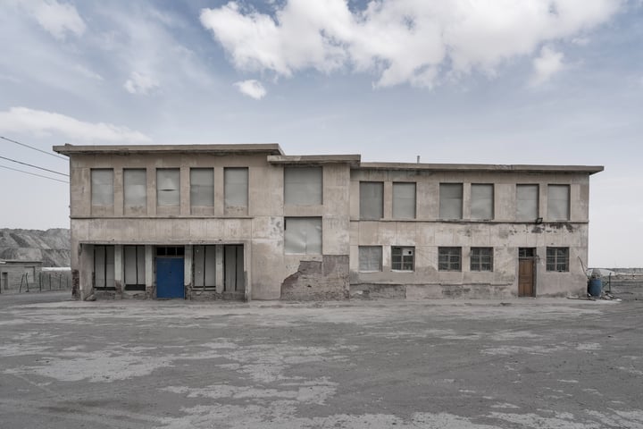 An old, dilapidated two-story building with a blue door and a large window on the second floor stands in a barren, empty lot. The sky is cloudy, and the ground is covered in dirt and gravel.