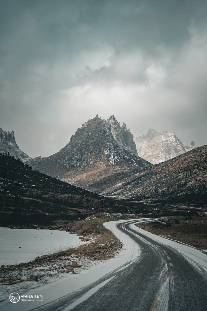 A snowy mountain landscape with a winding road, snow-covered mountains, and a cloudy sky.