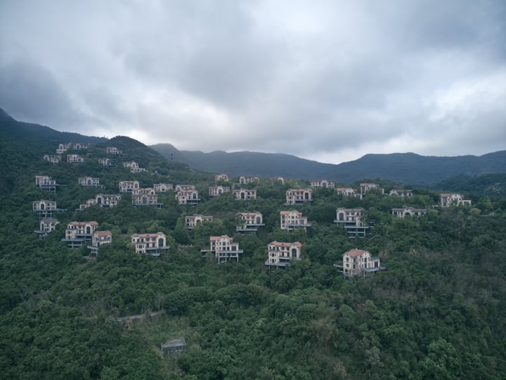 An aerial view of a densely forested hillside with numerous apartment buildings, some with balconies, arranged in a grid-like pattern. The buildings are predominantly white and gray, with some having red roofs. The hillside is covered in lush greenery, and the sky is cloudy.