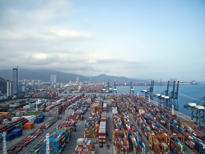 An aerial view of a bustling port with colorful shipping containers, cranes, and buildings, surrounded by mountains and water, showcasing a well-organized and efficient port.