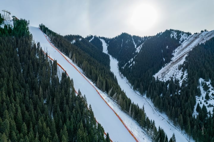 A snowy mountain slope with a red fence, surrounded by evergreen trees, is captured from a high vantage point, with sunlight shining through the trees.