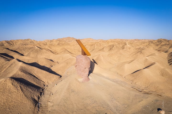 A large, pinkish-brown sand sculpture of a face with a yellow sun on top, standing on a hill in a desert landscape.