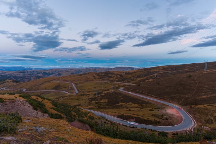 A winding, curvy road winds through a valley, surrounded by rolling hills and mountains, with power wind turbines in the distance.
