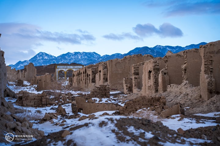 An old, weathered stone ruins stand in a barren landscape, with snow-covered mountains in the background. The sky is a deep blue with white clouds.