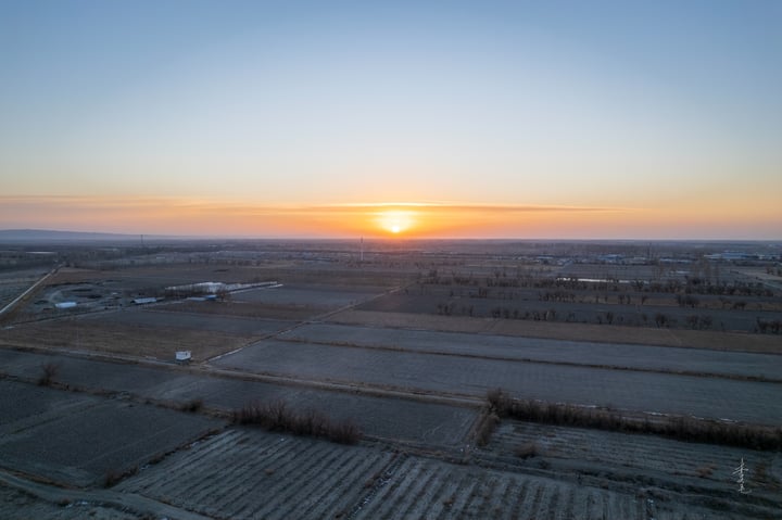 An aerial view of a vast, open field with a sun setting behind it, casting a warm glow on the landscape. Trees dot the field, and a small building is visible in the distance.