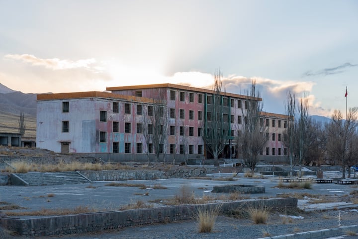 An old, dilapidated building with a pink facade and green roof stands in a barren, desolate landscape. The sky is filled with clouds, casting a soft glow on the scene.