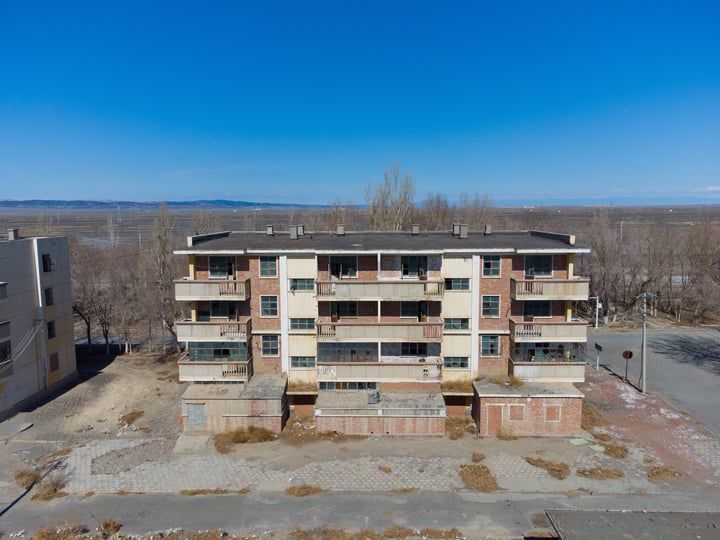 An abandoned apartment complex with multiple buildings, some with balconies, surrounded by a road and trees.