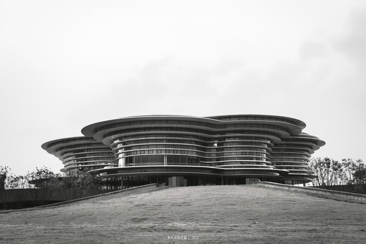 A black and white photograph captures a modern, curved building with numerous windows and a large, flat roof, situated on a grassy hill. The building is surrounded by trees and a few people are scattered around, adding life to the scene.
