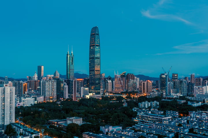 A cityscape at dusk features a tall, curved skyscraper with a pointed top, a modern office building, and a smaller, curved skyscraper. The city is densely populated with buildings, and the sky is a deep blue with a few clouds. The scene is illuminated by warm lights, creating a vibrant atmosphere.