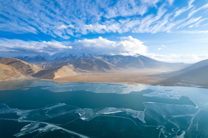 A serene landscape features a deep blue lake reflecting the sky, with snow-capped mountains in the distance. The sky is filled with white clouds, and the landscape is dominated by brown and beige tones.