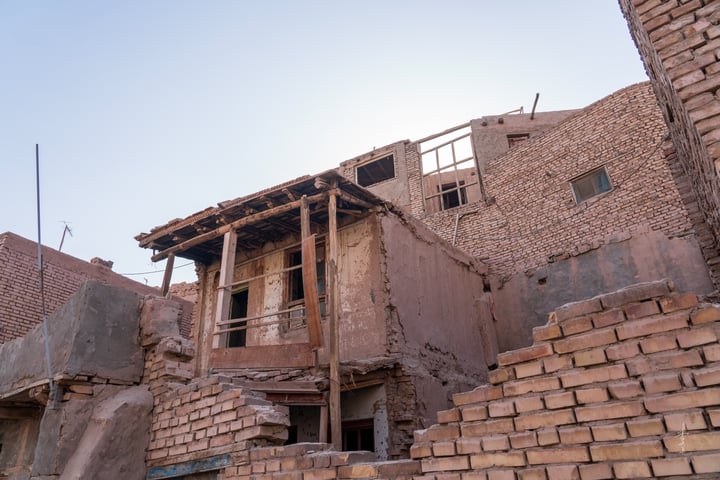 A dilapidated brick building with a wooden balcony and a collapsed roof, surrounded by a clear blue sky.