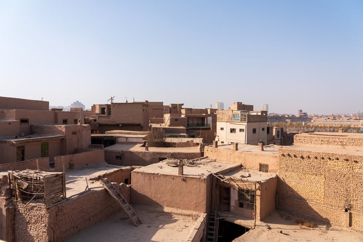 The image shows a desert cityscape with a mix of traditional and modern buildings, some with solar panels and others with traditional roofs. The city is surrounded by a hazy blue sky and a distant city skyline.