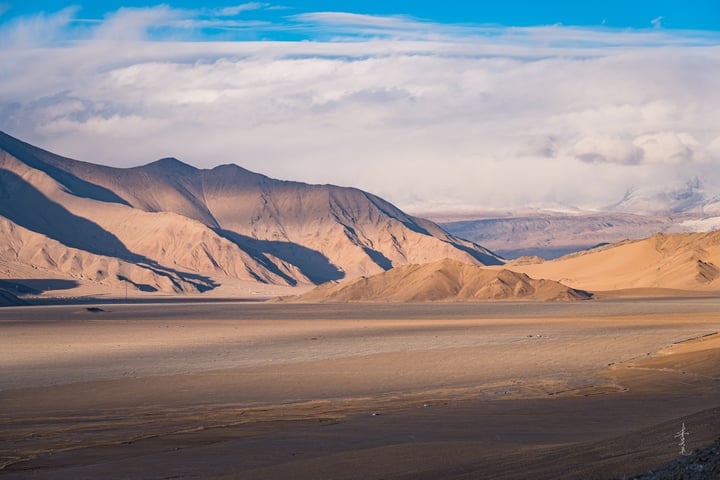 A vast desert landscape with brown sand and gray mountains, a blue sky with white clouds, and a distant white car on a dirt road.
