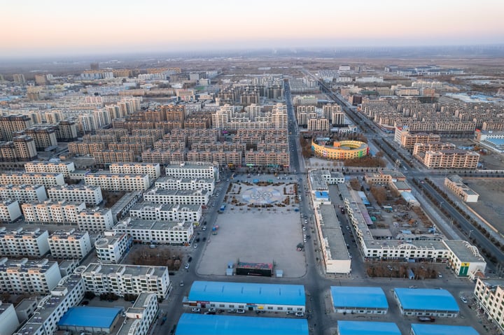 An aerial view of a bustling city with a large, yellow domed building in the center, surrounded by a sea of white and gray buildings. The city is filled with roads, roads, and buildings, and the sky is a pale blue.