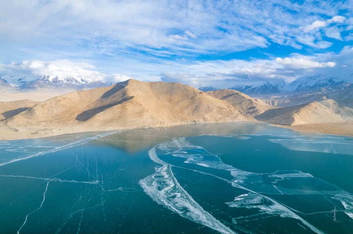 A serene landscape features a deep blue lake reflecting the sky, with snow-capped mountains in the distance. The sky is filled with white clouds, and the landscape is dominated by brown and beige tones.