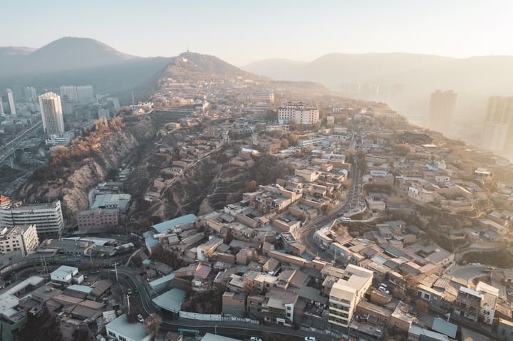 An aerial view of a mountainous city with a hillside, densely populated with buildings of varying heights, surrounded by greenery. The city is situated on a hill, with roads and highways running through it. The sun casts a warm glow on the city, highlighting its architectural details.
