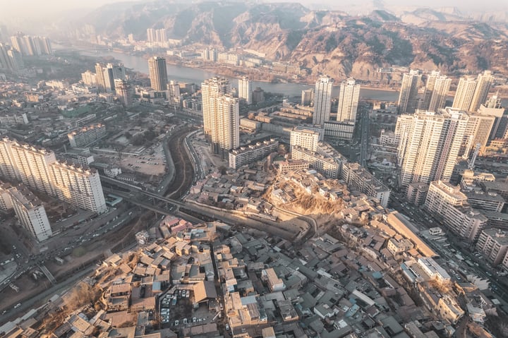 An aerial view of a mountainous city with a hillside, densely populated with buildings of varying heights, surrounded by greenery. The city is situated on a hill, with roads and highways running through it. The sun casts a warm glow on the city, highlighting its architectural details.