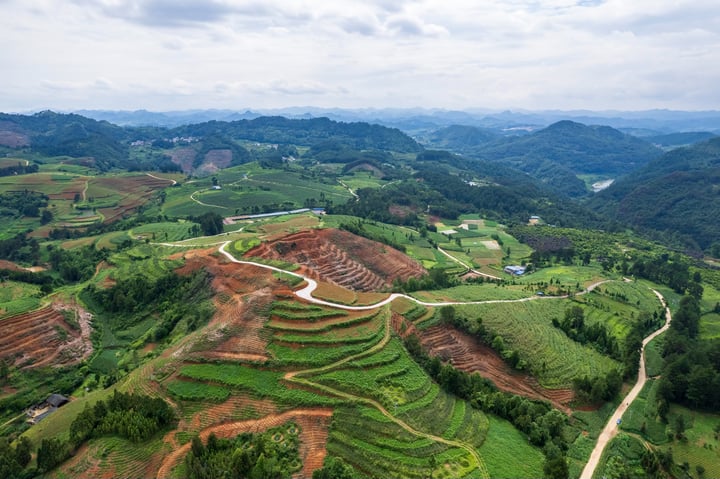 An aerial view of a mountainous landscape with terraced rice paddies, winding roads, and a blue sky with clouds.