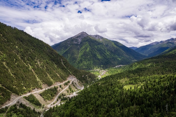 A winding, winding mountain road cuts through a lush valley, surrounded by towering mountains and a clear blue sky with white clouds.