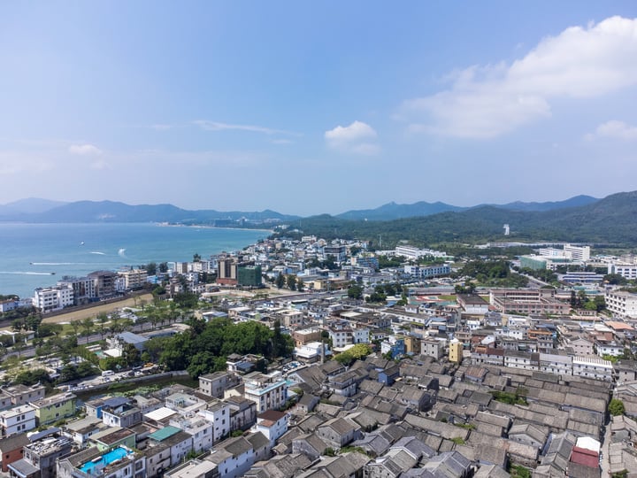 An aerial view of a city with a mix of white and gray buildings, surrounded by mountains and a large body of water.