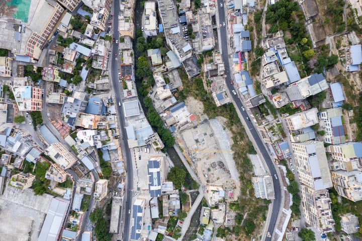 An aerial view of a city with a mix of residential and commercial buildings, surrounded by greenery and a road.
