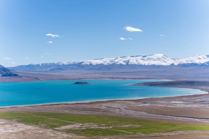A breathtaking aerial view of a vast, blue lake surrounded by snow-capped mountains, with a clear blue sky above.