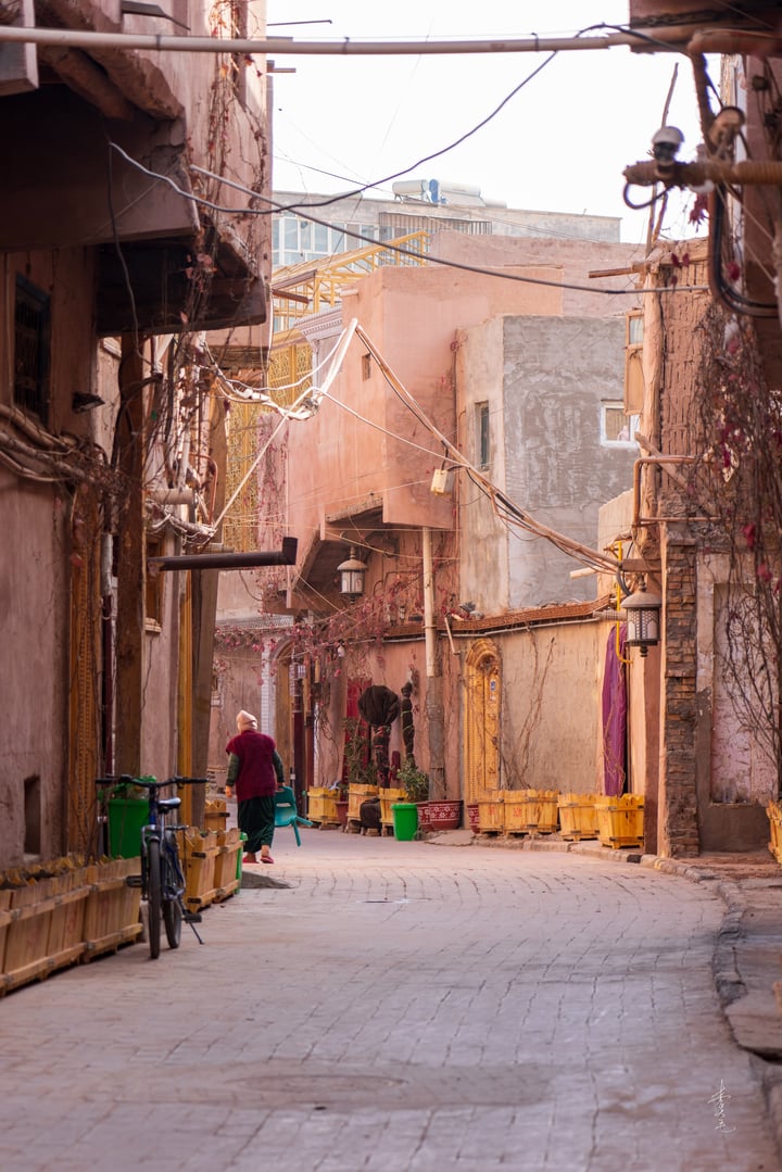 A narrow street in the Old City Kashgar, lined with old buildings and adorned with colorful decorations, is captured in a low-angle perspective. A man in a red shirt walks towards the camera, while a bicycle is parked on the left side. The scene is bathed in soft, diffused light, creating a serene atmosphere.