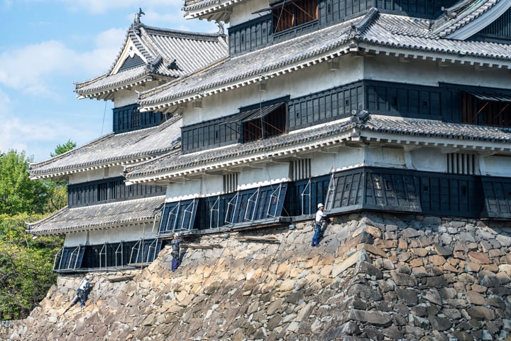 A traditional Japanese castle with a multi-level design, featuring black shutters and a gray roof, is perched on a stone wall. The castle is surrounded by lush green trees and a clear blue sky.
