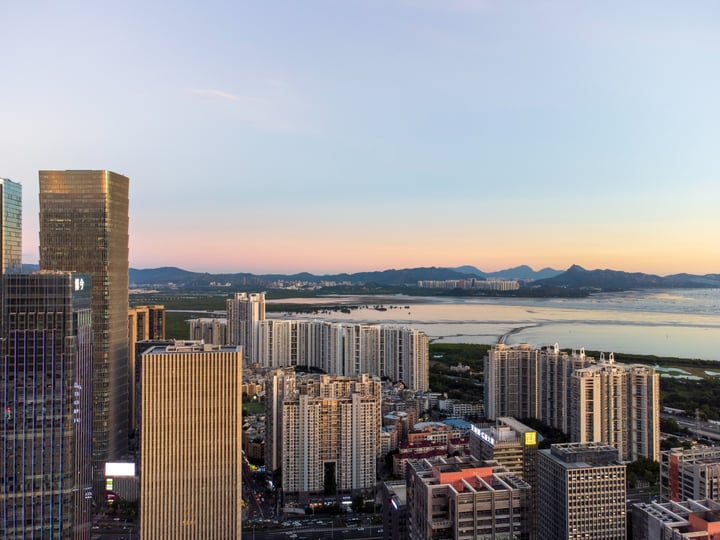 A cityscape at sunset, with tall buildings reflecting the setting sun, a calm body of water, and mountains in the distance.