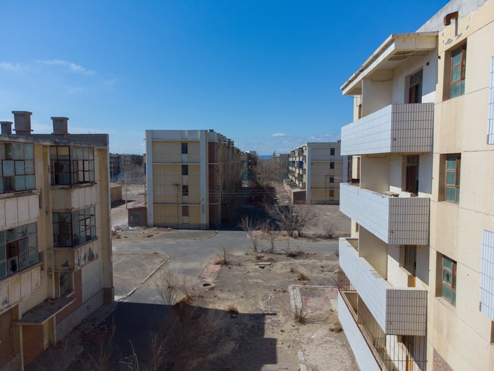 An old, abandoned cityscape with dilapidated buildings, a large empty lot, and a clear blue sky.