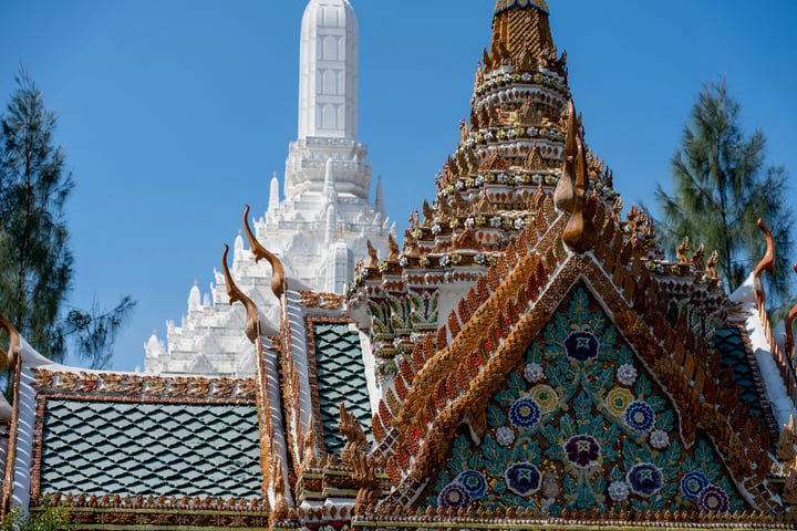 A majestic temple with a white spire and intricate gold and blue tilework stands tall against a clear blue sky, surrounded by lush green trees.